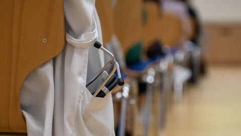 Nahaufnahme wie jemand auf einem Stuhl mit einem weißen Arztkittel sitzt. Der Kittel hängt an der Seite herunter und in der Tasche ist ein Stethoskop. 