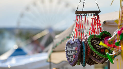 Lebkuchenherzen hängen von der Decke einer Bude. Im Hintergrund ist ein Riesenrad zu sehen.,