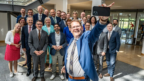 Über 20 Personen stehen dicht nebeneinander und posieren für ein Bild. Vorne steht ein Mann, der ein Handy in der Hand hält und von sich und den anderen ein Selfie macht.