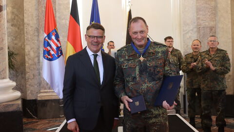 Ministerpräsident Boris Rhein steht neben Brigadegeneral Holger Radmann, der die Uniform der Bundeswehr trägt. Herr Radmann hält den Orden sowie die Urkunde in der Hand.