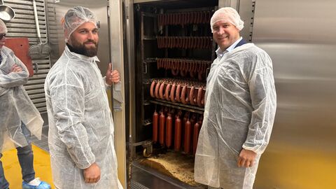 Zwei Personen stehen vor einem Räucherschrank und schauen in die Kamera.